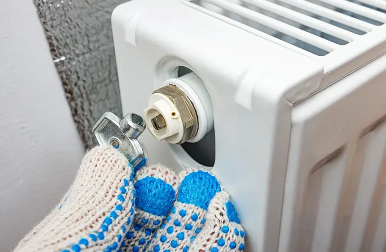 Gloved hands skillfully use a key to bleed a white radiator valve in Stoke-On-Trent. Positioned against the wall, the key turns to release trapped air, enhancing heating efficiency.