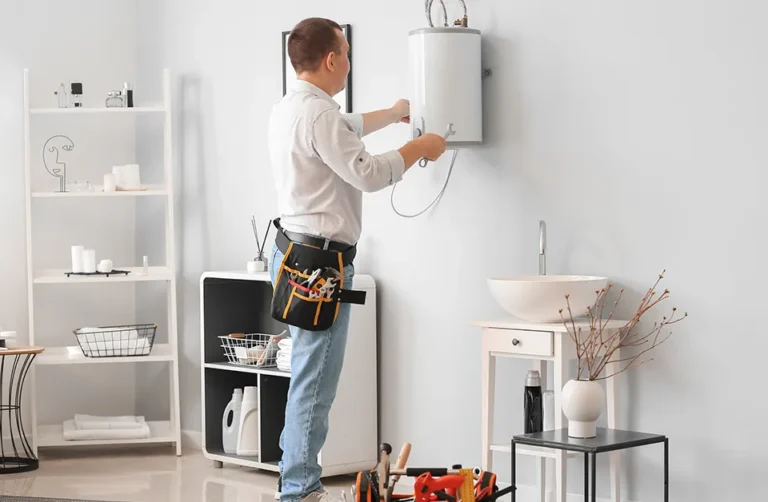 A man in jeans and a white shirt is busy with a boiler service in Stoke-on-Trent, carefully fixing a white water heater on the gray wall. With a tool belt around his waist, shelves and a sink nearby, and a toolbox on the floor, he works efficiently to get the job done.