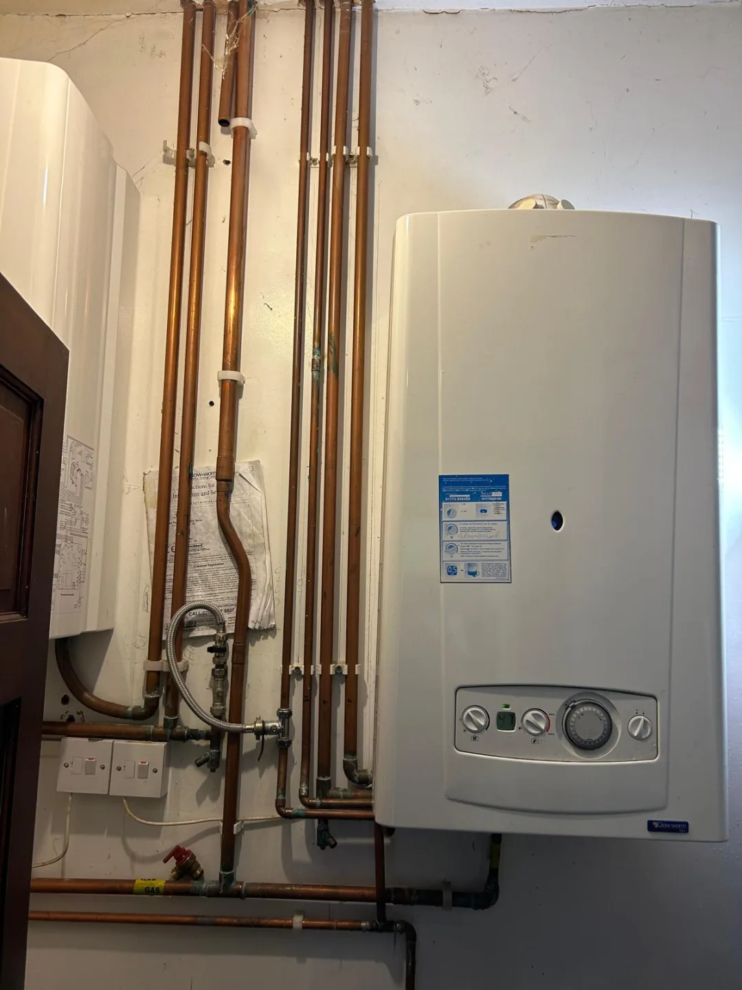 Wall-mounted combi boiler beside copper pipework and a hot water cylinder in a domestic cupboard