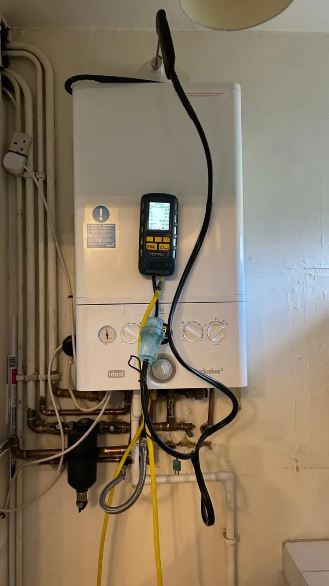 Gas analyser hanging from a wall-mounted boiler during a flue gas test, pipework and valves below